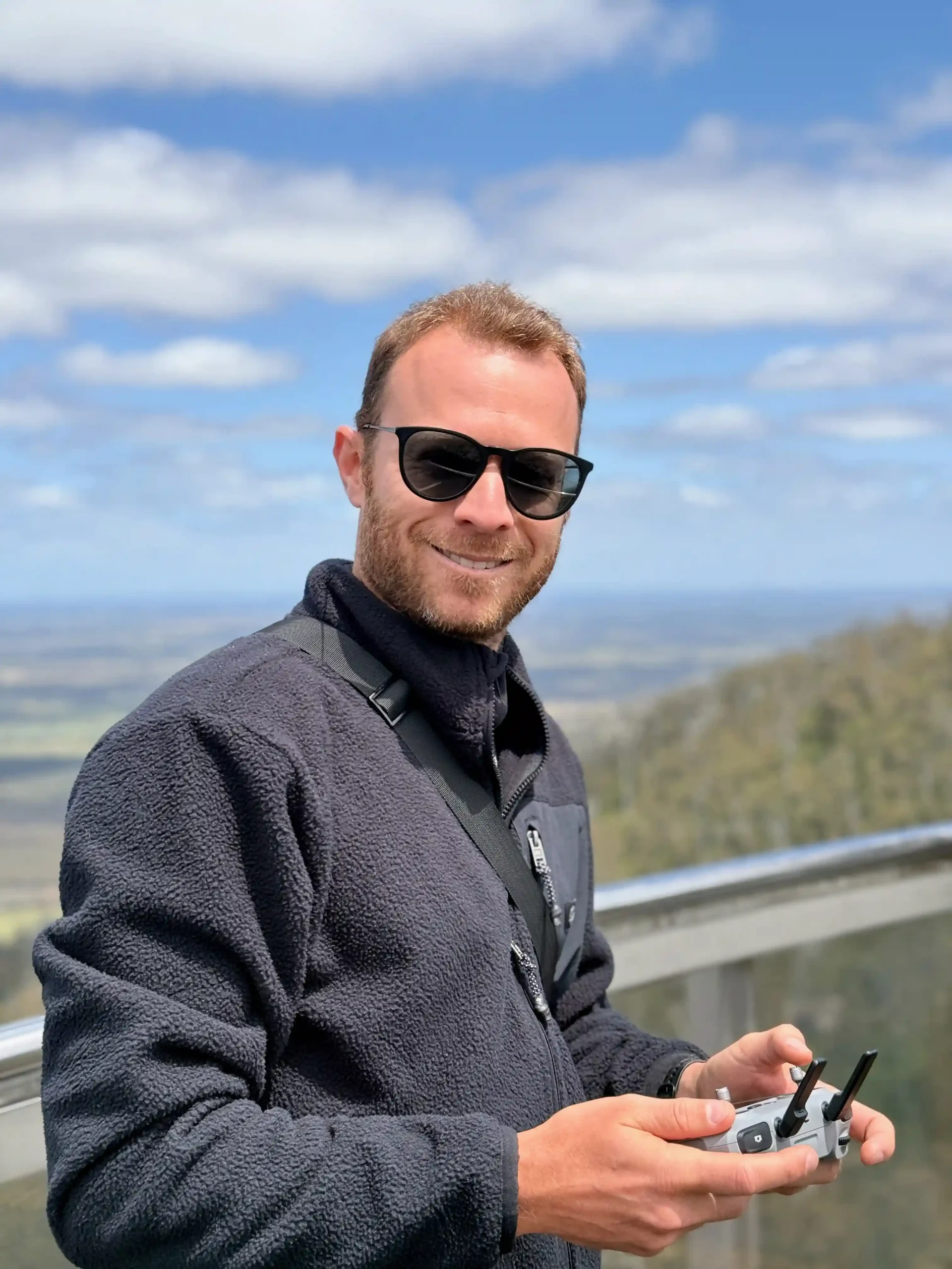 Alt text: A person wearing a navy fleece jacket and black sunglasses smiles while holding a drone controller outdoors. The photo is taken from a high vantage point with a scenic landscape and blue sky with white clouds visible in the background.